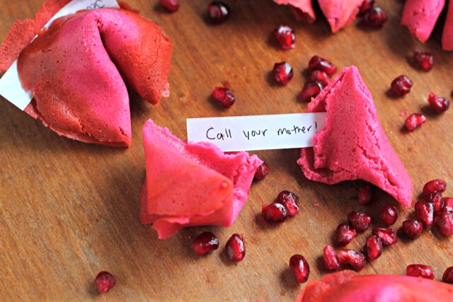 Pink pomegranate fortune cookies on a wooden board with pomegranate seeds and a fortune reading Call your mother